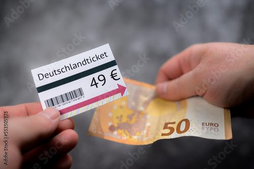 Hands holding 49-Euro-Ticket mockup and 50 euro cash bill against out of focus gray background with barcode and minimal design elements on the ticket visible photo