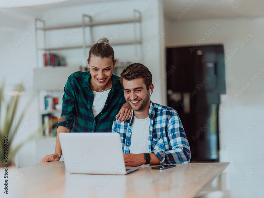 A young married couple is talking to parents, family and friends on a video call via a laptop while sitting in the living room of their modern house A young married couple is talking to parents, family and friends on a video call via a laptop while sitting in the living room of their modern house