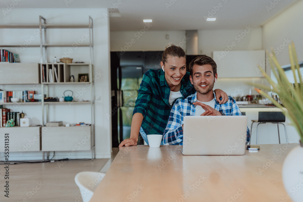 Obraz premium A young married couple is talking to parents, family and friends on a video call via a laptop while sitting in the living room of their modern house