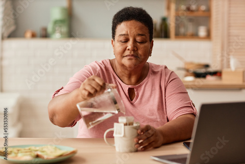 Warm toned portrait of black senior woman making tea during breakfast in cozy kitchen
