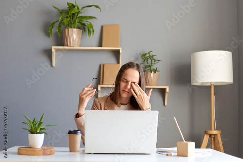 Indoor shot of tired woman wearing official style jacket sitting in office at desk in front of laptop, rubbing her eyes, feels pain, looking at computer display long hours.