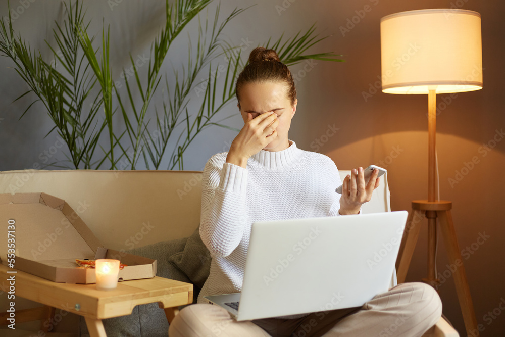 Φωτογραφία Portrait of exhausted woman with bun hairstyle wearing white sweater sitting on cough and working on laptop, feels pain in her eyes, working long hours