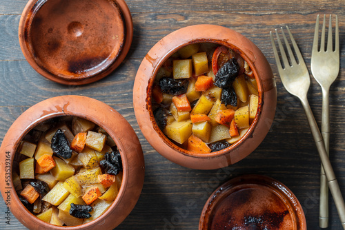Stewed potato, carrot, onion, tomato and prunes in a clay pots with lid on wooden background, closeup