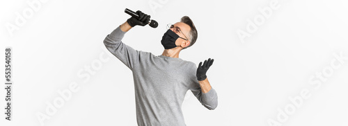 Concept of coronavirus, lifestyle and quarantine. Portrait of attractive middle-aged man in black medical mask and gloves, singing in microphone, standing over white background