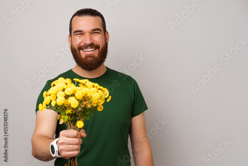 Handsome bearded man is holding a bunch of yellow flowers giving the to the camera.