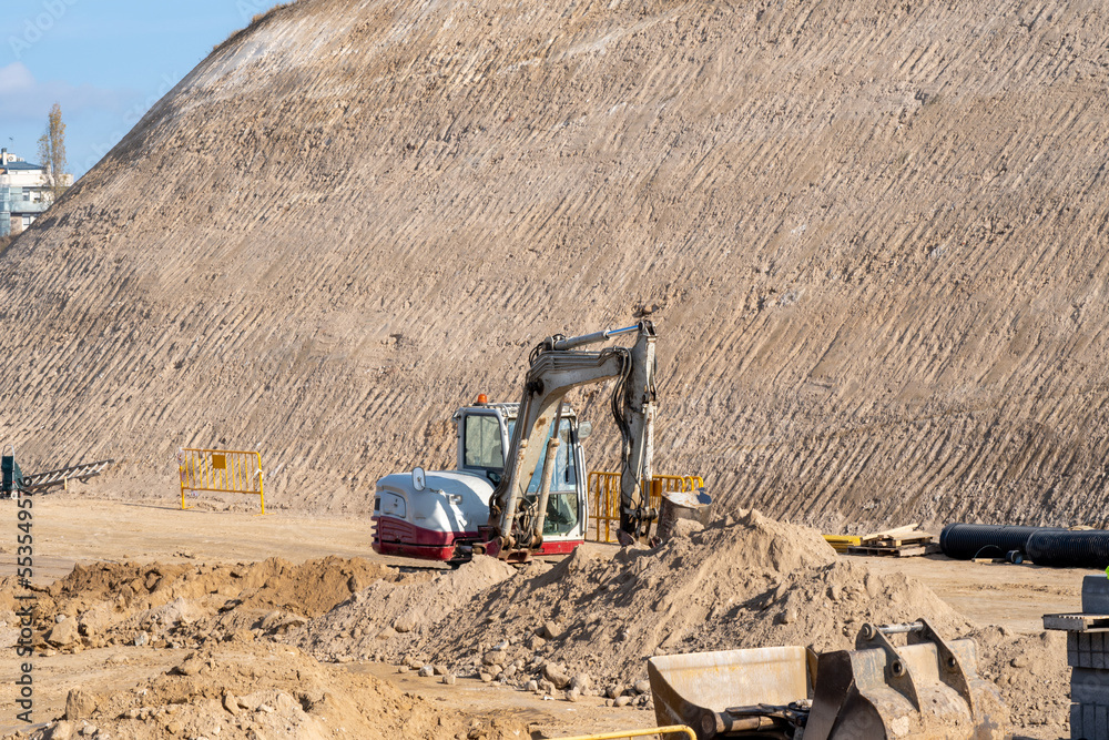 White and red small excavator moving earth and sand in the middle of a large construction site