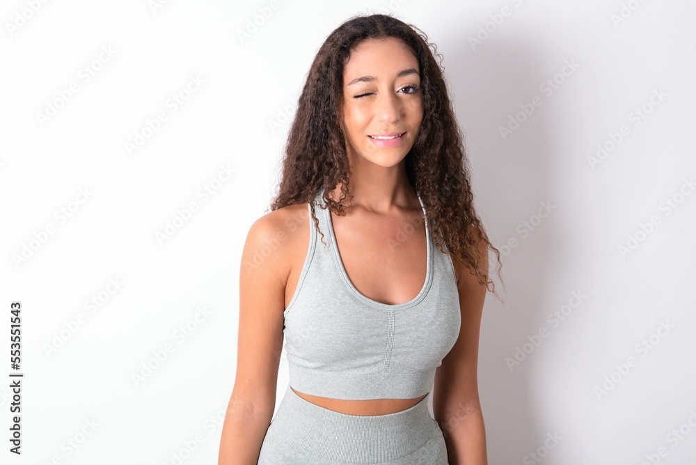 Coquettish teen girl with curly hair wearing grey sport set over white background smiling happily, blinking at camera in a playful manner, flirting with you. Coquettish teen girl with curly hair wearing grey sport set over white background smiling happily, blinking at camera in a playful manner, flirting with you.