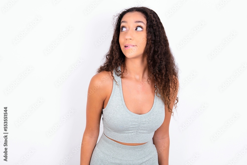 Amazed teen girl with curly hair wearing grey sport set over white background bitting lip and looking tricky to empty space. Amazed teen girl with curly hair wearing grey sport set over white background bitting lip and looking tricky to empty space.