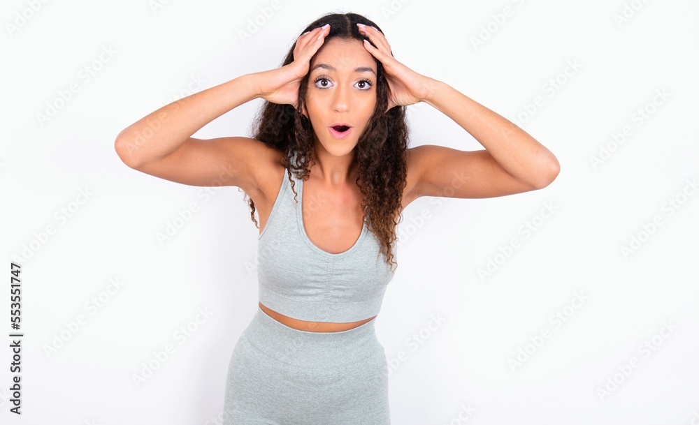 teen girl with curly hair wearing grey sport set over white background with scared expression, keeps hands on head, jaw dropped, has terrific expression. Omg concept teen girl with curly hair wearing grey sport set over white background with scared expression, keeps hands on head, jaw dropped, has terrific expression. Omg concept