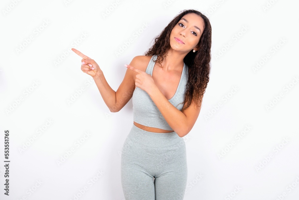 teen girl with curly hair wearing grey sport set over white background indicating finger empty space showing best low prices, looking at the camera teen girl with curly hair wearing grey sport set over white background indicating finger empty space showing best low prices, looking at the camera