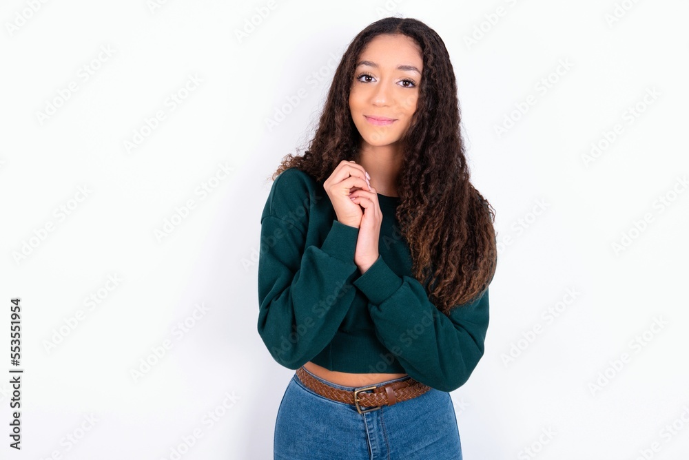 Dreamy charming teen girl with curly hair wearing green sweater over white background with pleasant expression, keeps hands crossed near face, excited about something pleasant. Dreamy charming teen girl with curly hair wearing green sweater over white background with pleasant expression, keeps hands crossed near face, excited about something pleasant.