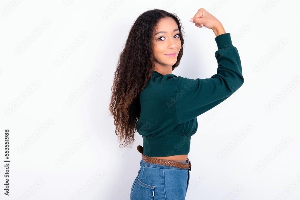 teen girl with curly hair wearing green sweater over white background, showing muscles after workout. Health and strength concept. teen girl with curly hair wearing green sweater over white background, showing muscles after workout. Health and strength concept.