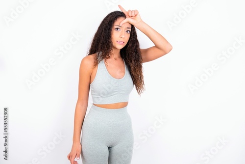 teen girl with curly hair wearing white sport set over gray background making fun of people with fingers on forehead doing loser gesture mocking and insulting.