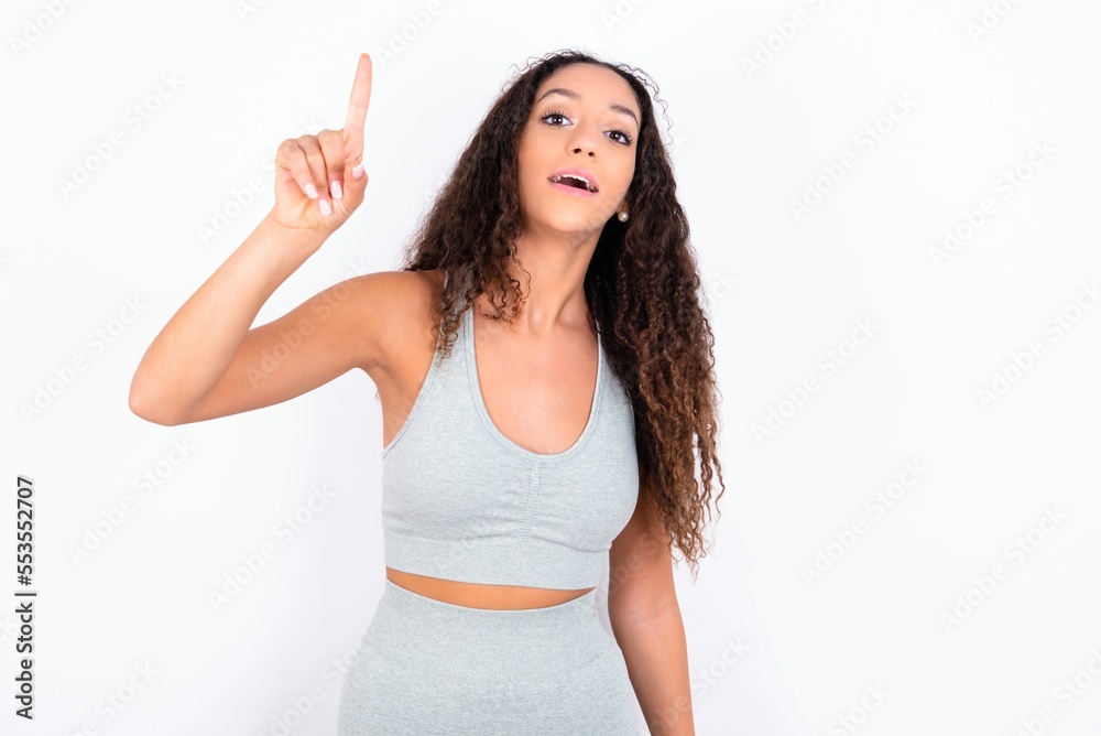 Beautiful teen girl with curly hair wearing grey sport set over white background pointing finger up and looking inspired by genius thought, showing good idea sign, having clever solution in mind Stock-Foto | Beautiful teen girl with curly hair wearing grey sport set over white background pointing finger up and looking inspired by genius thought, showing good idea sign, having clever solution in mind Stock-Foto |