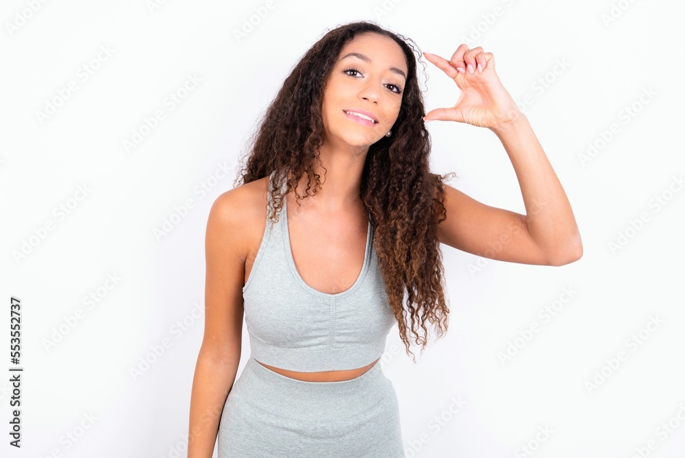 Beautiful teen girl with curly hair wearing grey sport set over white background smiling and confident gesturing with hand doing small size sign with fingers looking and the camera. Measure concept Beautiful teen girl with curly hair wearing grey sport set over white background smiling and confident gesturing with hand doing small size sign with fingers looking and the camera. Measure concept