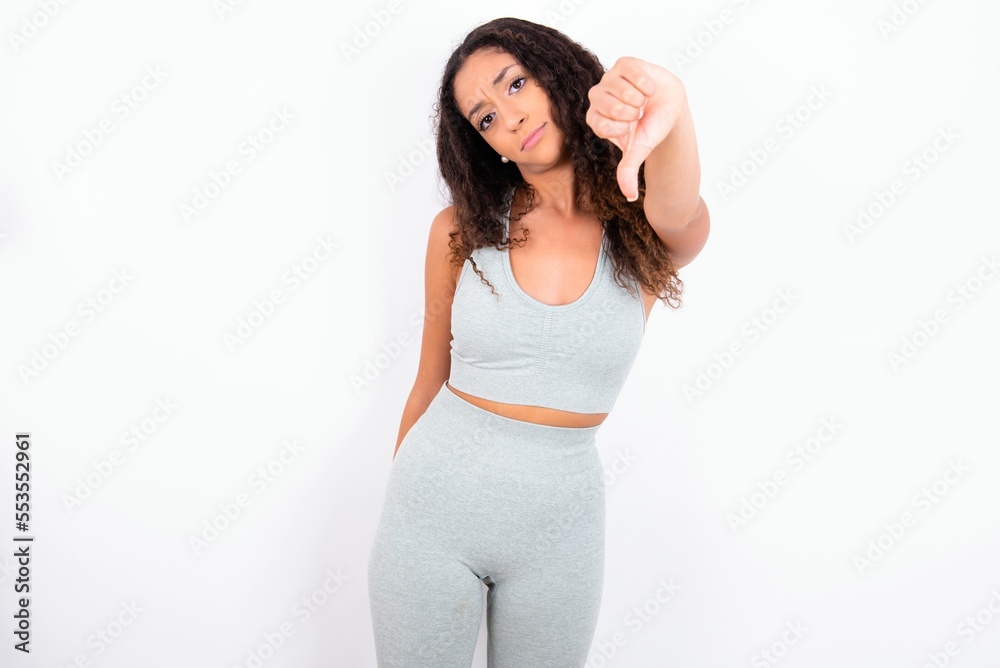 Beautiful teen girl with curly hair wearing grey sport set over white background feeling angry, annoyed, disappointed or displeased, showing thumbs down with a serious look Beautiful teen girl with curly hair wearing grey sport set over white background feeling angry, annoyed, disappointed or displeased, showing thumbs down with a serious look