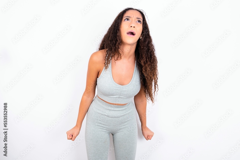 Beautiful teen girl with curly hair wearing grey sport set over white background angry and mad screaming frustrated and furious, shouting with anger. Rage and aggressive concept. Beautiful teen girl with curly hair wearing grey sport set over white background angry and mad screaming frustrated and furious, shouting with anger. Rage and aggressive concept.