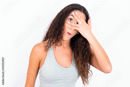 Beautiful teen girl with curly hair wearing grey sport set over white background peeking in shock covering face and eyes with hand, looking through fingers with embarrassed expression.