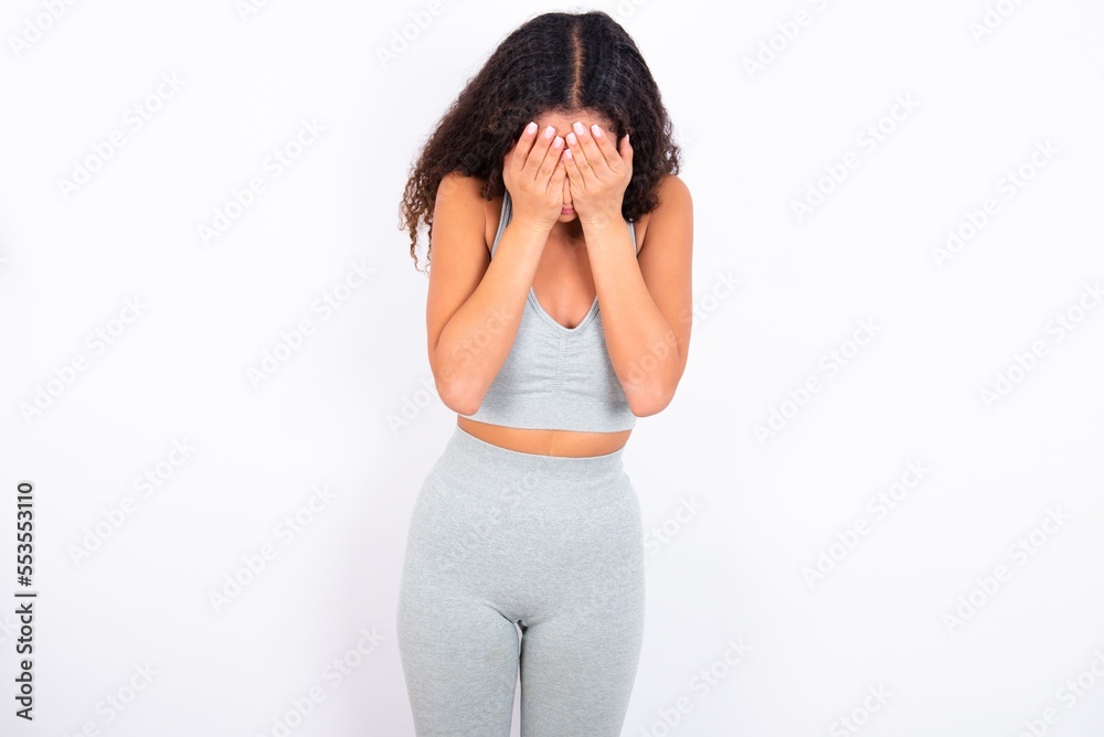 Beautiful teen girl with curly hair wearing grey sport set over white background covering her face with her hands, being devastated and crying. Sad concept