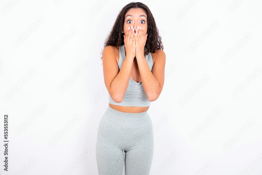 Vivacious Beautiful teen girl with curly hair wearing grey sport set over white background, giggles joyfully, covers mouth, has natural laughter, hears positive story or funny anecdote