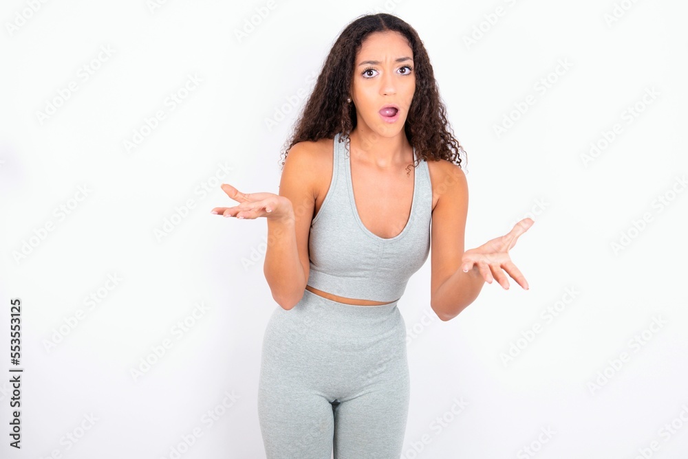 Frustrated Beautiful teen girl with curly hair wearing grey sport set over white wall feels puzzled and hesitant, shrugs shoulders in bewilderment, keeps mouth widely opened, doesn't know what to do. Frustrated Beautiful teen girl with curly hair wearing grey sport set over white wall feels puzzled and hesitant, shrugs shoulders in bewilderment, keeps mouth widely opened, doesn't know what to do.