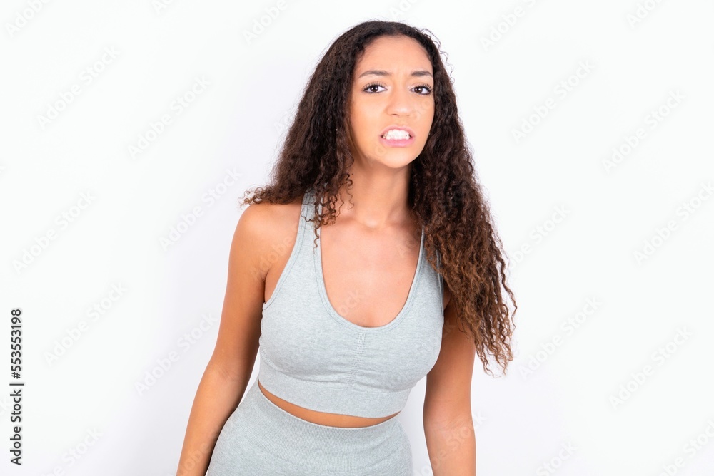 Beautiful teen girl with curly hair wearing green sport set over white background keeps teeth clenched, frowns face in dissatisfaction, irritated because of much duties. Beautiful teen girl with curly hair wearing green sport set over white background keeps teeth clenched, frowns face in dissatisfaction, irritated because of much duties.