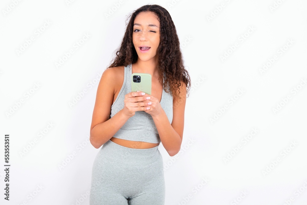 Beautiful teen girl with curly hair wearing green sport set over white background taking a selfie celebrating success Beautiful teen girl with curly hair wearing green sport set over white background taking a selfie celebrating success