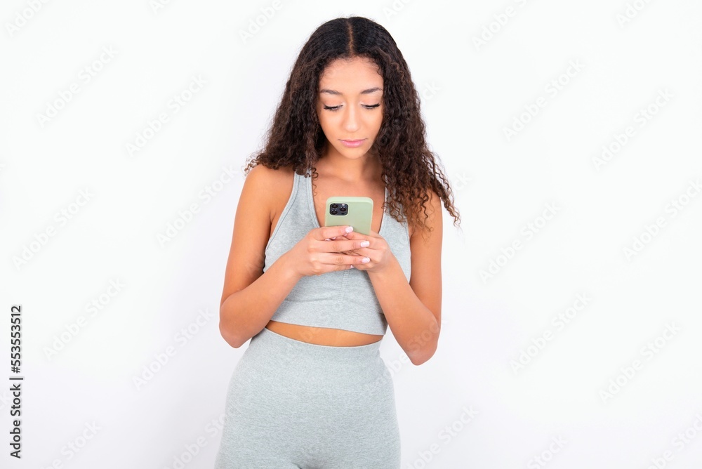 Excited Beautiful teen girl with curly hair wearing green sport set over white background winking and eye hold smart phone use read social network news Excited Beautiful teen girl with curly hair wearing green sport set over white background winking and eye hold smart phone use read social network news