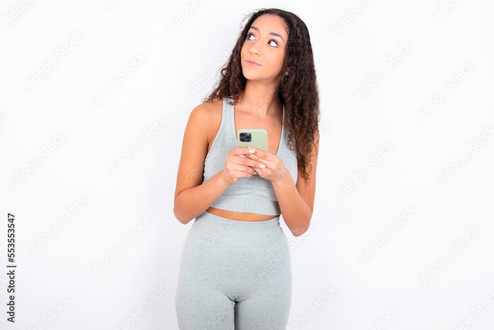 Beautiful teen girl with curly hair wearing green sport set over white background holds telephone hands reads good youth news look empty space advert Beautiful teen girl with curly hair wearing green sport set over white background holds telephone hands reads good youth news look empty space advert