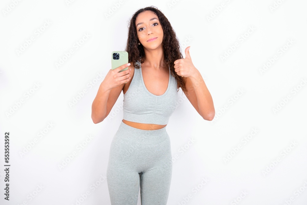 Portrait of Beautiful teen girl with curly hair wearing green sport set over white wall using and texting with smartphone happy with big smile doing ok sign, thumb up with fingers, excellent sign Portrait of Beautiful teen girl with curly hair wearing green sport set over white wall using and texting with smartphone happy with big smile doing ok sign, thumb up with fingers, excellent sign