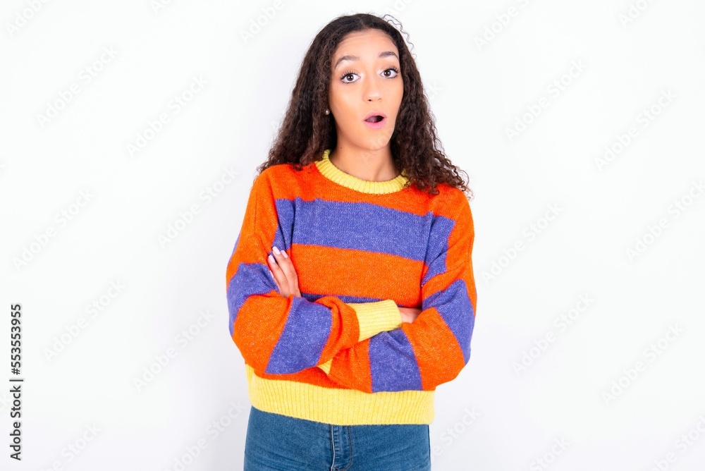 Shocked embarrassed beautiful teen girl wearing striped knitted sweater over white background keeps mouth widely opened. Hears unbelievable novelty stares in stupor Shocked embarrassed beautiful teen girl wearing striped knitted sweater over white background keeps mouth widely opened. Hears unbelievable novelty stares in stupor