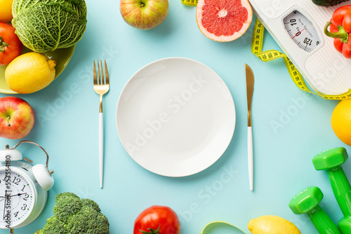 Proper nutrition concept. Top view photo of plate fork knife scales vegetables fruits alarm clock dumbbells and tape measure on isolated pastel blue background with empty space