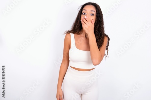 teen girl with curly hair wearing white sport set over white background covers mouth and looks with wonder at camera, cannot believe unexpected rumors.