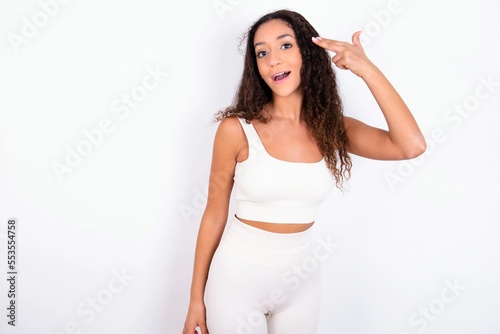 teen girl with curly hair wearing white sport set over white background foolishness around shoots in temple with fingers makes suicide gesture. Funny model makes finger gun pistol photo