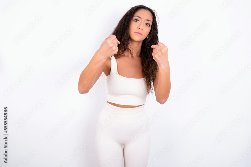 Displeased annoyed teen girl with curly hair wearing white sport set over white clenches fists, gestures pissed, ready to revenge, looks with aggression at camera stands full of hate, being pressured Displeased annoyed teen girl with curly hair wearing white sport set over white clenches fists, gestures pissed, ready to revenge, looks with aggression at camera stands full of hate, being pressured