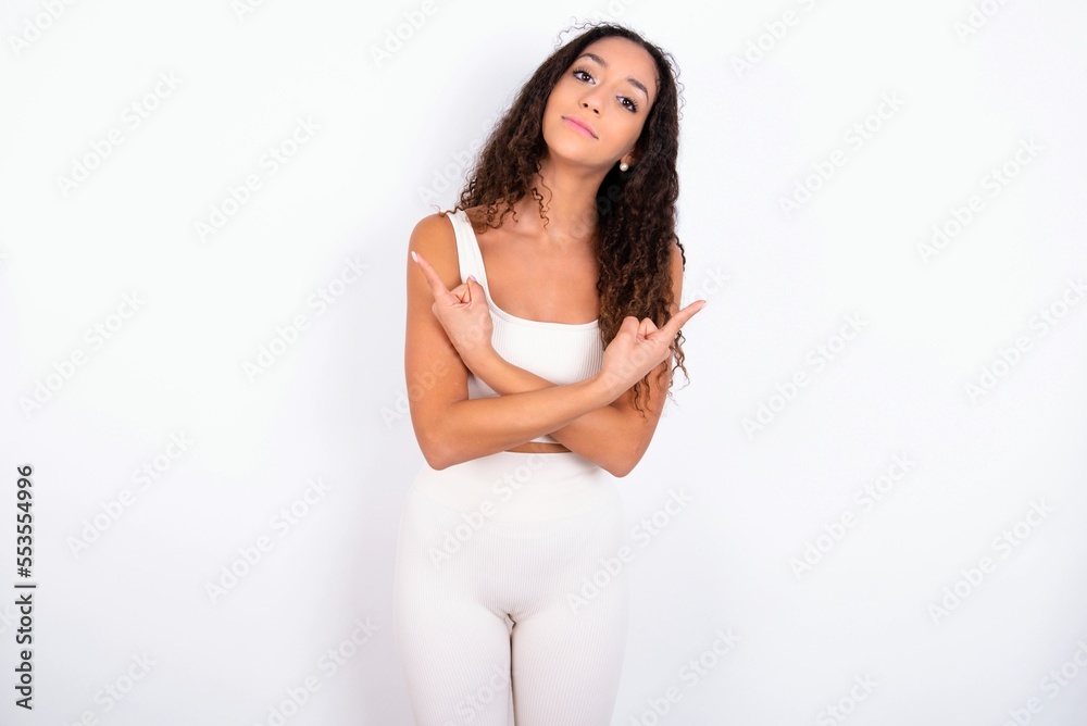 teen girl with curly hair wearing white sport set over white background crosses arms and points at different sides hesitates between two items or variants. Needs help with decision teen girl with curly hair wearing white sport set over white background crosses arms and points at different sides hesitates between two items or variants. Needs help with decision