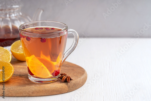 Healthy vitamin hot tea drink served in transparent glass cup made with cranberries, lemon citrus fruit slices and sweet honey on white wooden table table with teapot, anise. Image with copy space