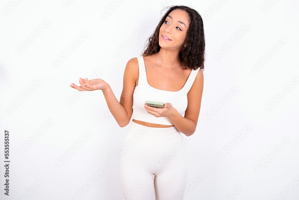 Happy pleased teen girl with curly hair wearing white sport set over white background raises palm and holds cellphone uses high speed internet for text messaging or video calls Happy pleased teen girl with curly hair wearing white sport set over white background raises palm and holds cellphone uses high speed internet for text messaging or video calls