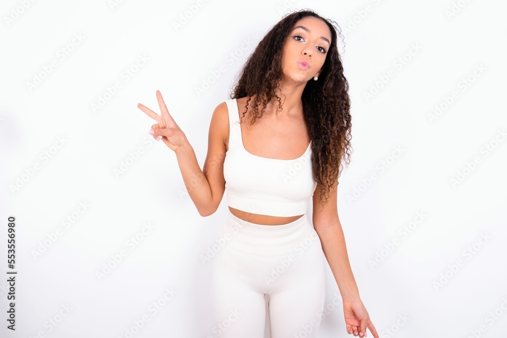teen girl with curly hair wearing white sport set over white background makes peace gesture keeps lips folded shows v sign. Body language concept teen girl with curly hair wearing white sport set over white background makes peace gesture keeps lips folded shows v sign. Body language concept
