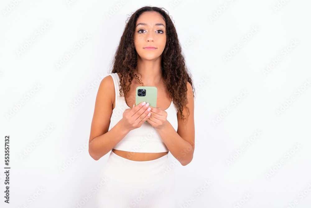 Portrait of serious confident teen girl with curly hair wearing white sport set over white background holding phone in two hands Portrait of serious confident teen girl with curly hair wearing white sport set over white background holding phone in two hands