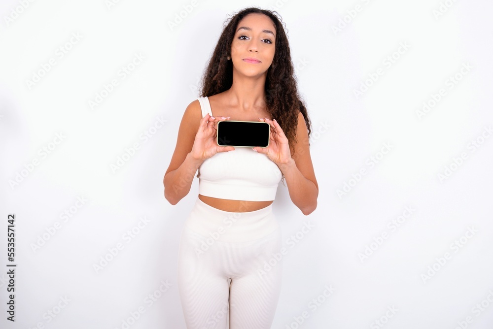 Cheerful cheery content teen girl with curly hair wearing white sport set over white background holding in hands device hobby smm post blog Cheerful cheery content teen girl with curly hair wearing white sport set over white background holding in hands device hobby smm post blog