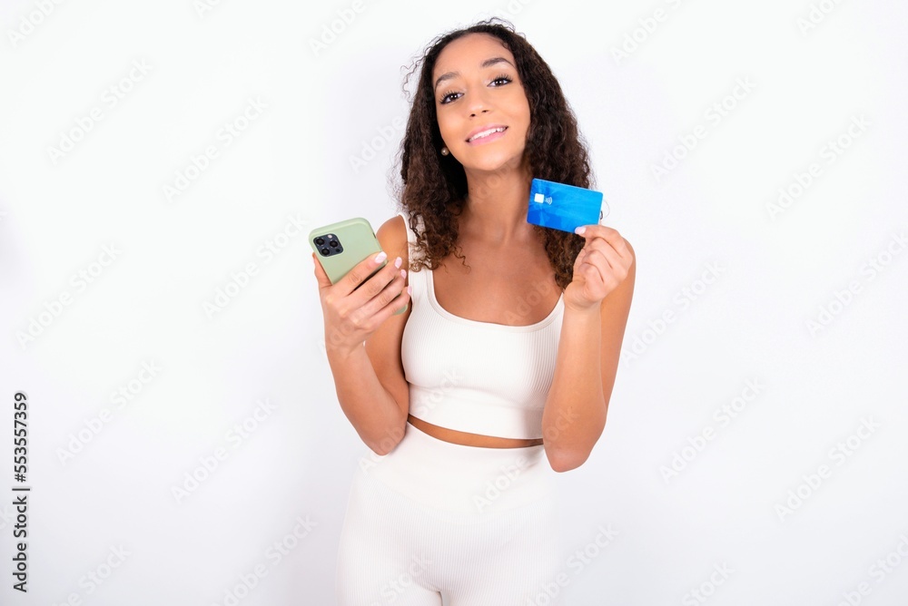 teen girl with curly hair wearing white sport set over white background wears stereo headphones on ears holds modern mobile phone and credit card teen girl with curly hair wearing white sport set over white background wears stereo headphones on ears holds modern mobile phone and credit card