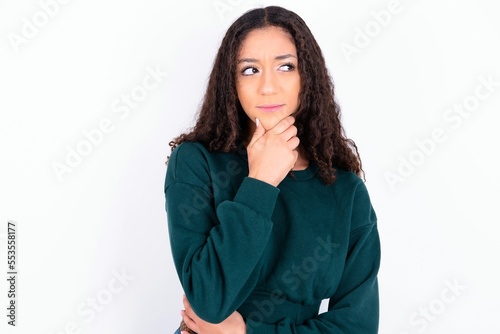 Shot of contemplative thoughtful teen girl wearing knitted green sweater over white background keeps hand under chin, looks thoughtfully upwards. © Jihan
