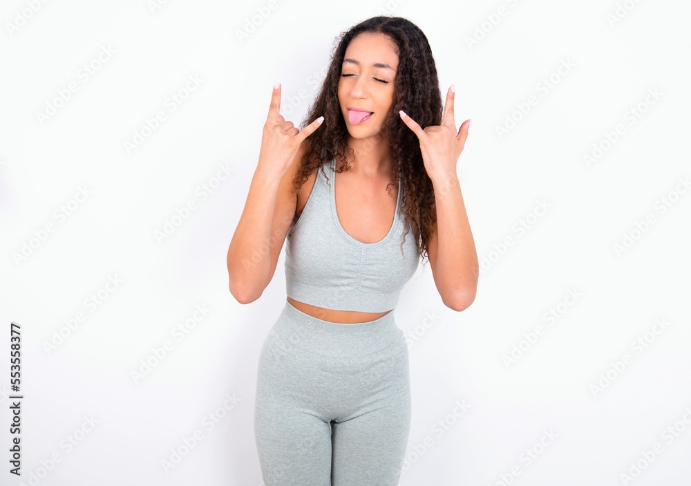 teen girl with curly hair wearing white sport set over gray background making rock hand gesture and showing tongue teen girl with curly hair wearing white sport set over gray background making rock hand gesture and showing tongue