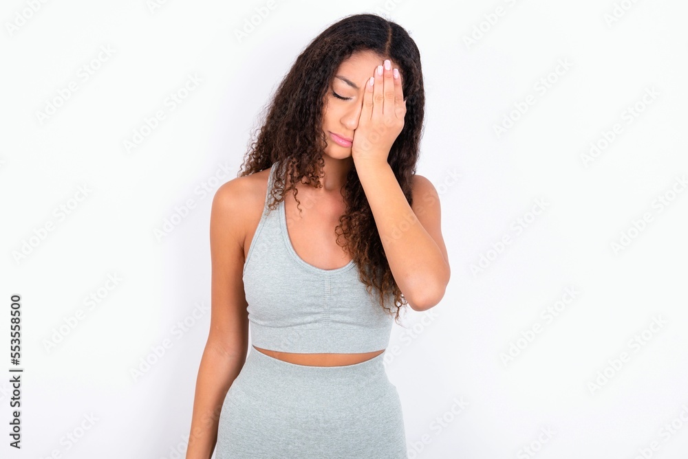 Tired overworked teen girl with curly hair wearing white sport set over gray wall has sleepy expression, gloomy look, covers face with hand, has eyes shut, gasps from tiredness, fatigue after party Tired overworked teen girl with curly hair wearing white sport set over gray wall has sleepy expression, gloomy look, covers face with hand, has eyes shut, gasps from tiredness, fatigue after party