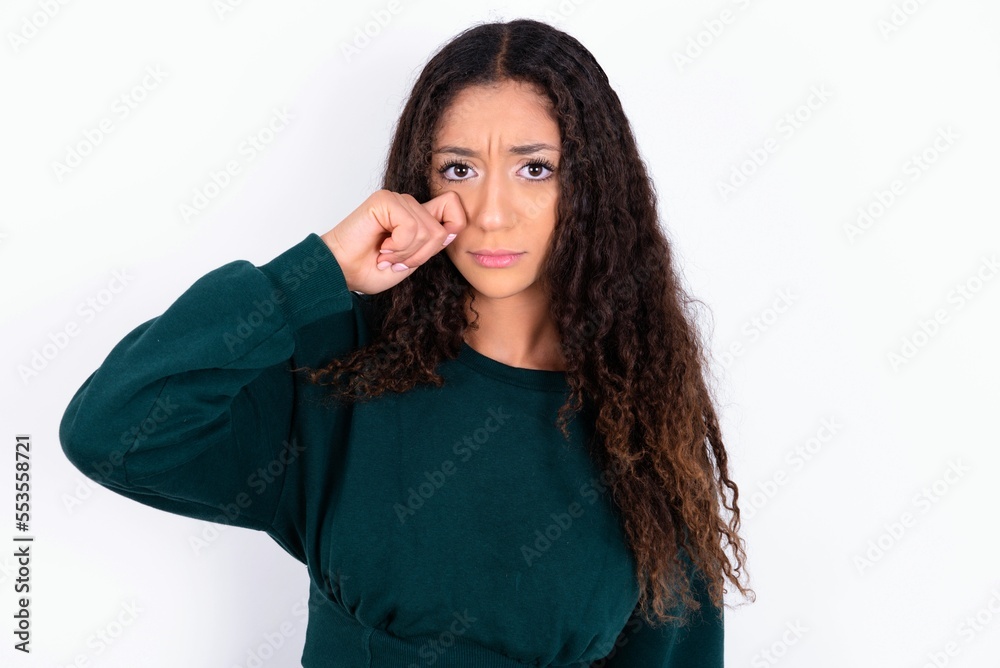 Unhappy teen girl wearing knitted green sweater over white background crying while posing at camera whipping tears with hand. Unhappy teen girl wearing knitted green sweater over white background crying while posing at camera whipping tears with hand.