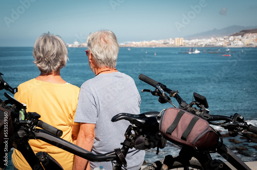 Back view of relaxed senior couple in vacation or retirement at sea enjoying healthy lifestyle with bicycles. Sitting outdoors looking at the horizon over water