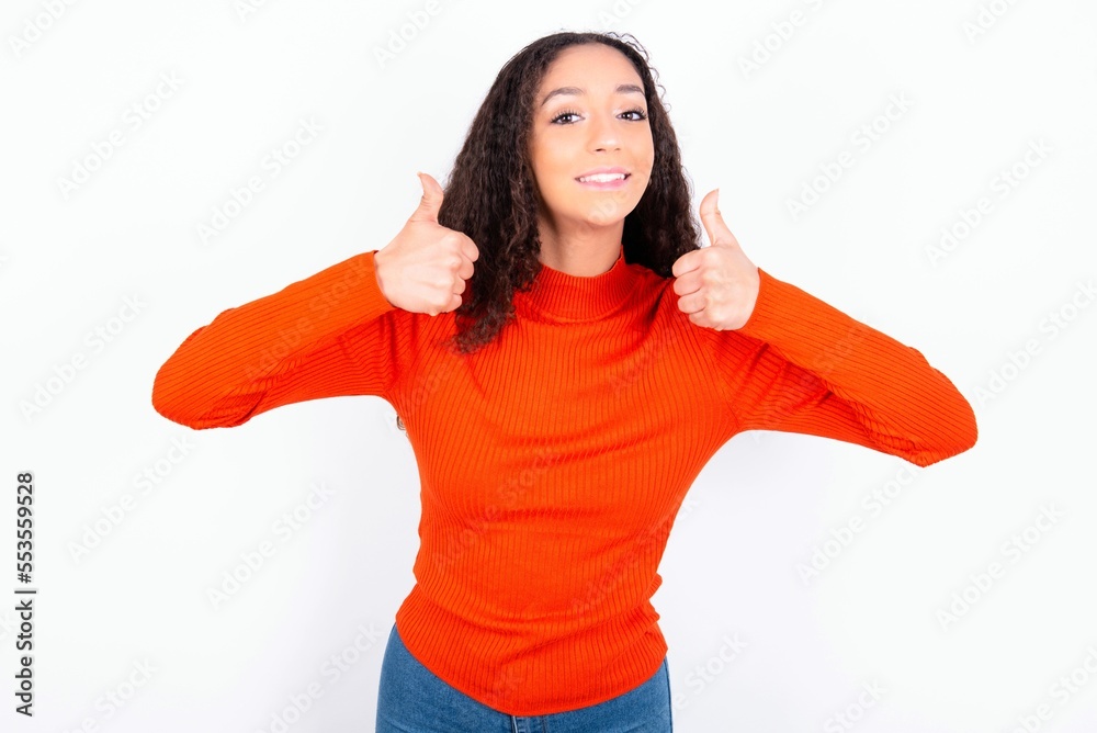 Optimistic teen girl wearing knitted red sweater over white background showing thumbs up with positive emotions. Quality and recommendation concept. Optimistic teen girl wearing knitted red sweater over white background showing thumbs up with positive emotions. Quality and recommendation concept.
