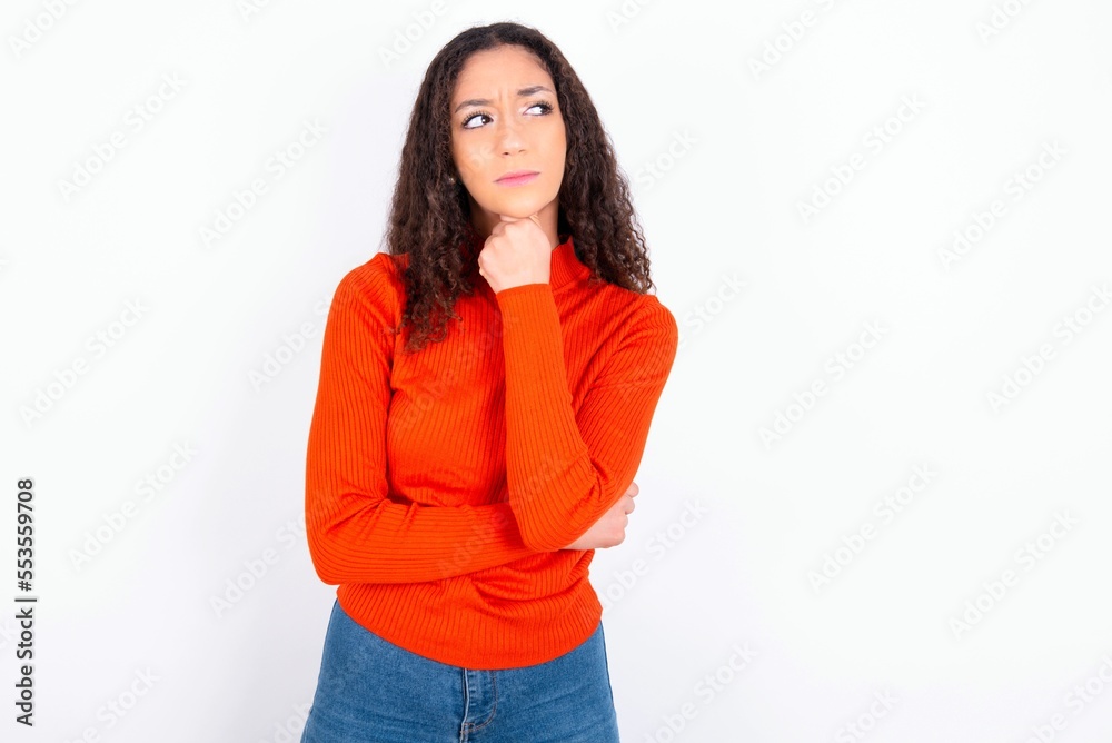 teen girl wearing knitted red sweater over white background with hand under chin and looking sideways with doubtful and skeptical expression, suspect and doubt. teen girl wearing knitted red sweater over white background with hand under chin and looking sideways with doubtful and skeptical expression, suspect and doubt.