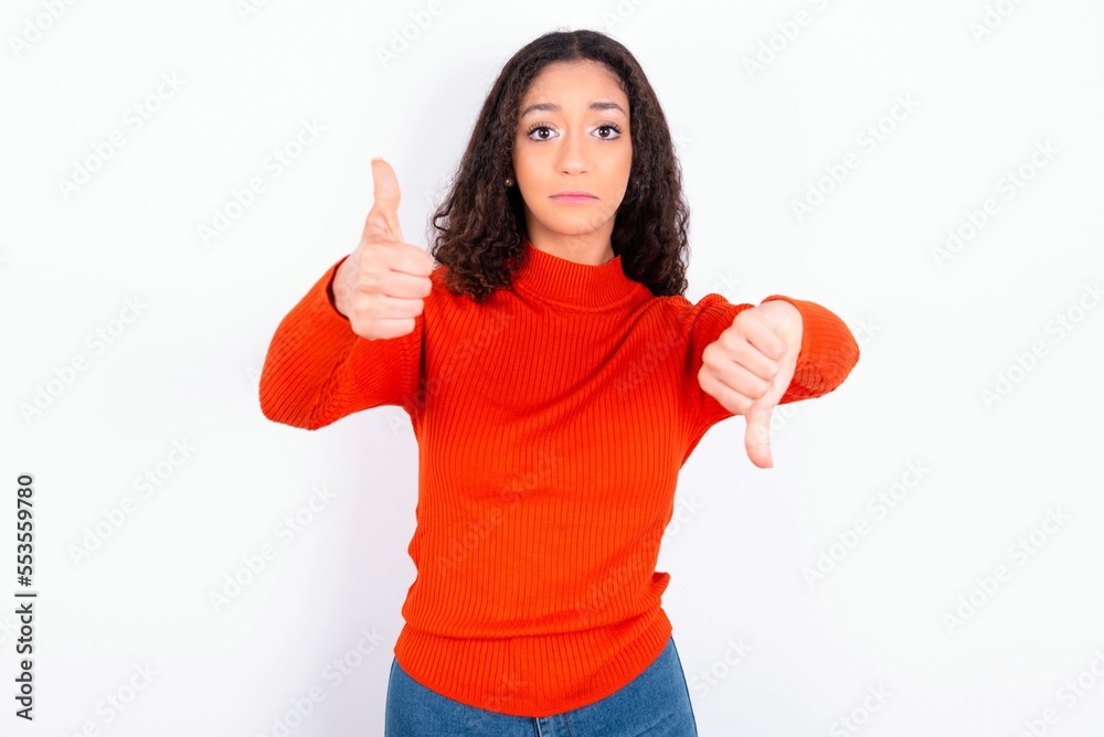 teen girl wearing knitted red sweater over white background feeling unsure making good bad sign. Displeased and unimpressed. teen girl wearing knitted red sweater over white background feeling unsure making good bad sign. Displeased and unimpressed.